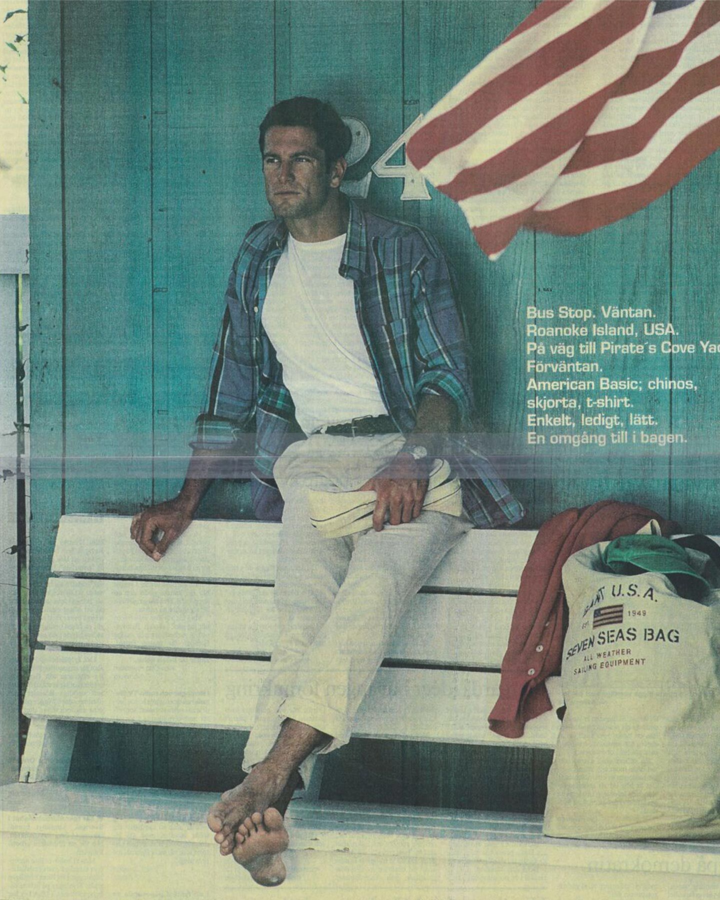 A man in Gant chinos and a white shirt sits at an old bus stop with a “SEVEN SEAS” bag and a U.S. flag behind him.
